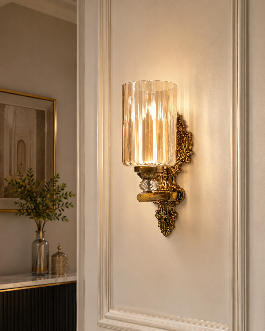 An ornate brass wall sconce with a clear ribbed glass cylinder shade, mounted on a white paneled wall in an elegant interior setting.