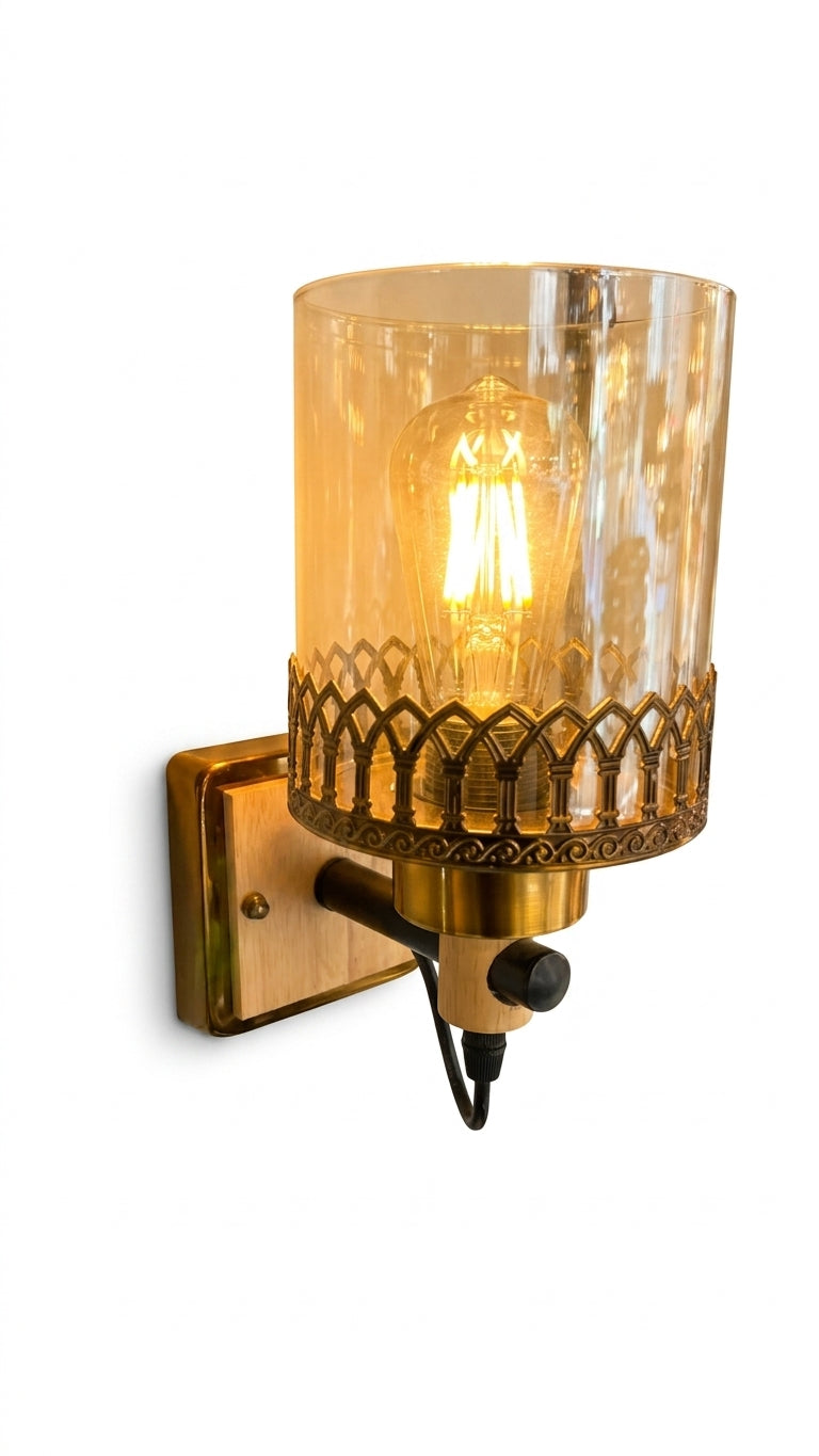 A wall-mounted light fixture with a cylindrical amber-tinted glass shade, an ornate gold-toned decorative band, and a visible glowing filament bulb. The fixture is attached to a square wall plate with a black power cord hanging below, shown against a plain white background.