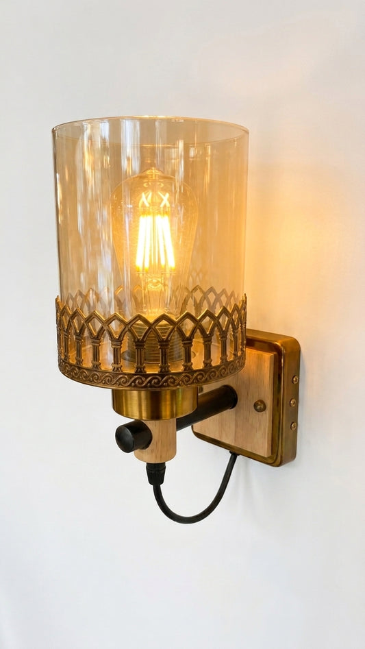 A close-up studio photograph of an elegant wall sconce featuring a brushed brass C-shaped arm that supports a delicate crystal prism and a cylindrical