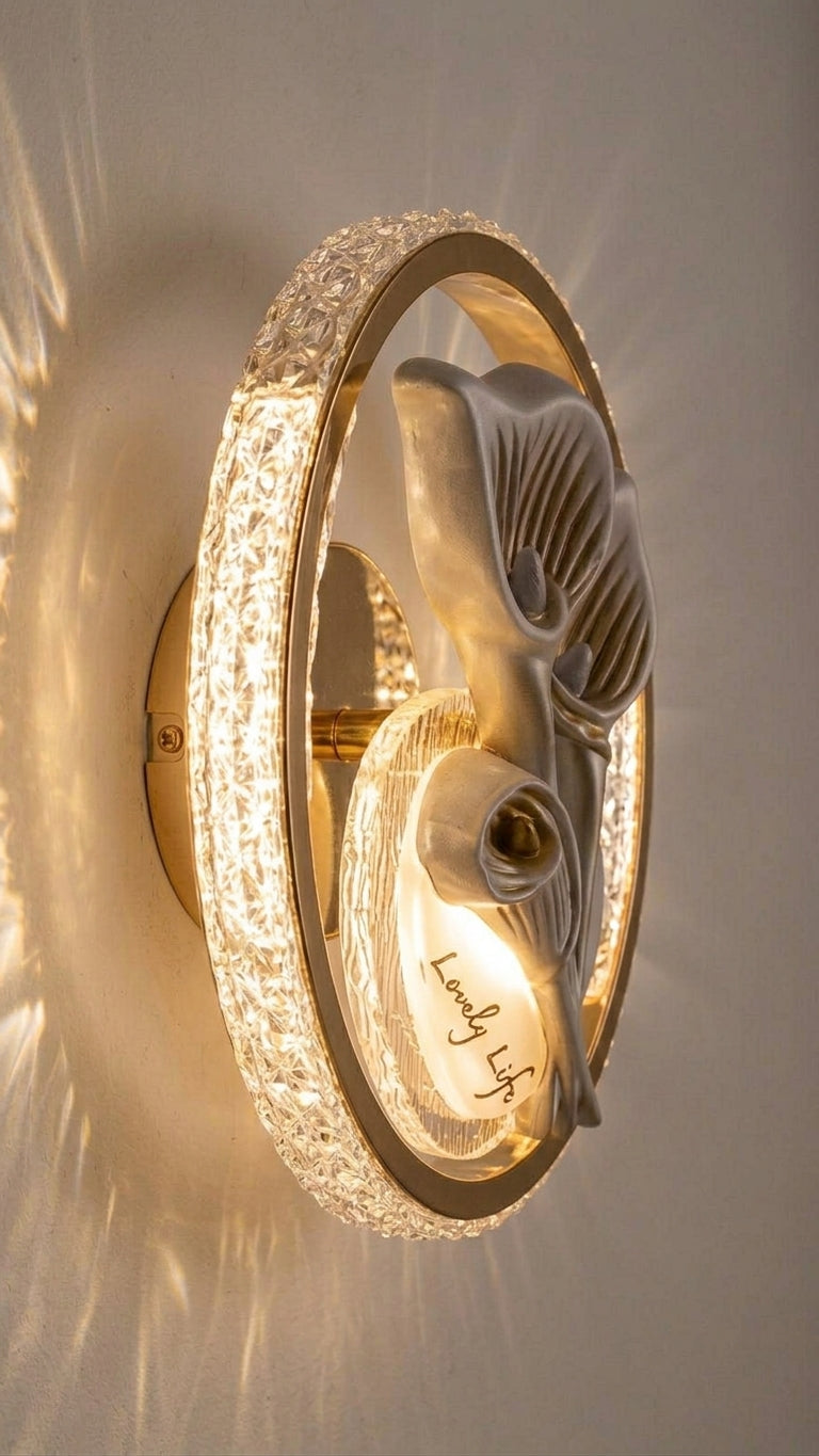 A modern wall-mounted light fixture with a circular clear crystal ring and a sculptural white floral or leaf-like ornament in the center, shown lit against a neutral wall.