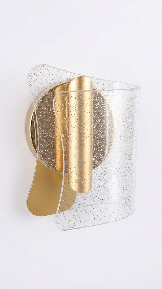 A modern wall sconce light fixture with a brushed gold metal body and a clear, curved glass shade filled with fine silver glitter, mounted on a plain white wall.