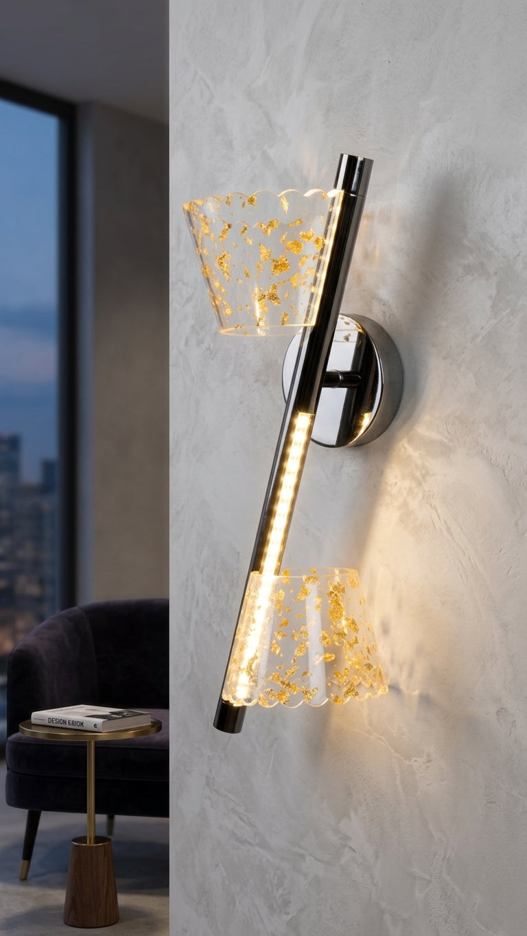 A modern wall-mounted sconce with a vertical brushed metal stem, two clear glass shades with gold leaf flecks, and an integrated warm white LED light, shown mounted on a textured light gray wall in a styled interior setting.