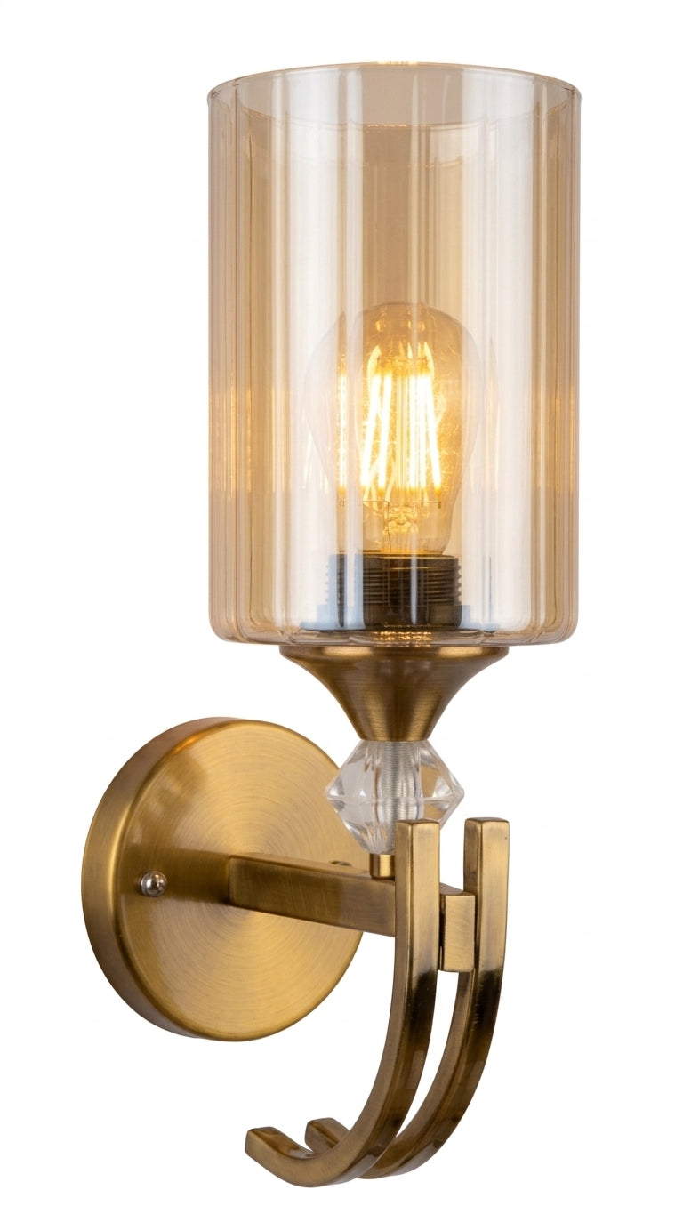 A wall-mounted sconce light fixture with a brushed brass/gold metal wall plate and curved bracket, featuring a clear ribbed glass cylinder shade, a visible warm Edison-style bulb, and a small faceted crystal detail.