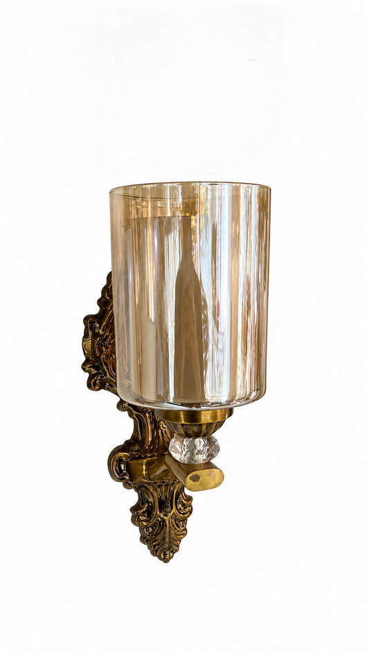 An ornate brass wall sconce with a clear ribbed glass cylinder shade and a clear crystal accent, shown against a plain white background.
