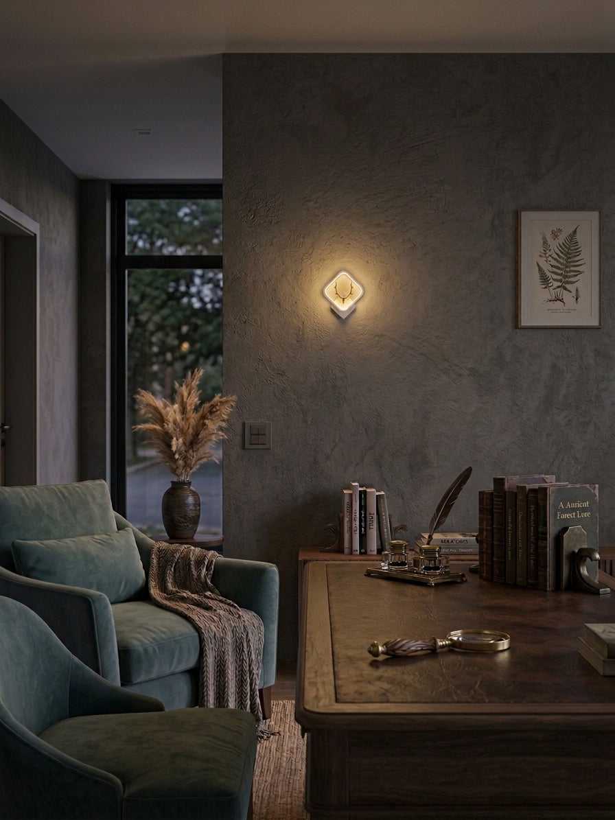 A modern diamond-shaped wall sconce with a warm glowing light fixture, mounted on a textured dark gray wall in a cozy, dimly lit living room setting.