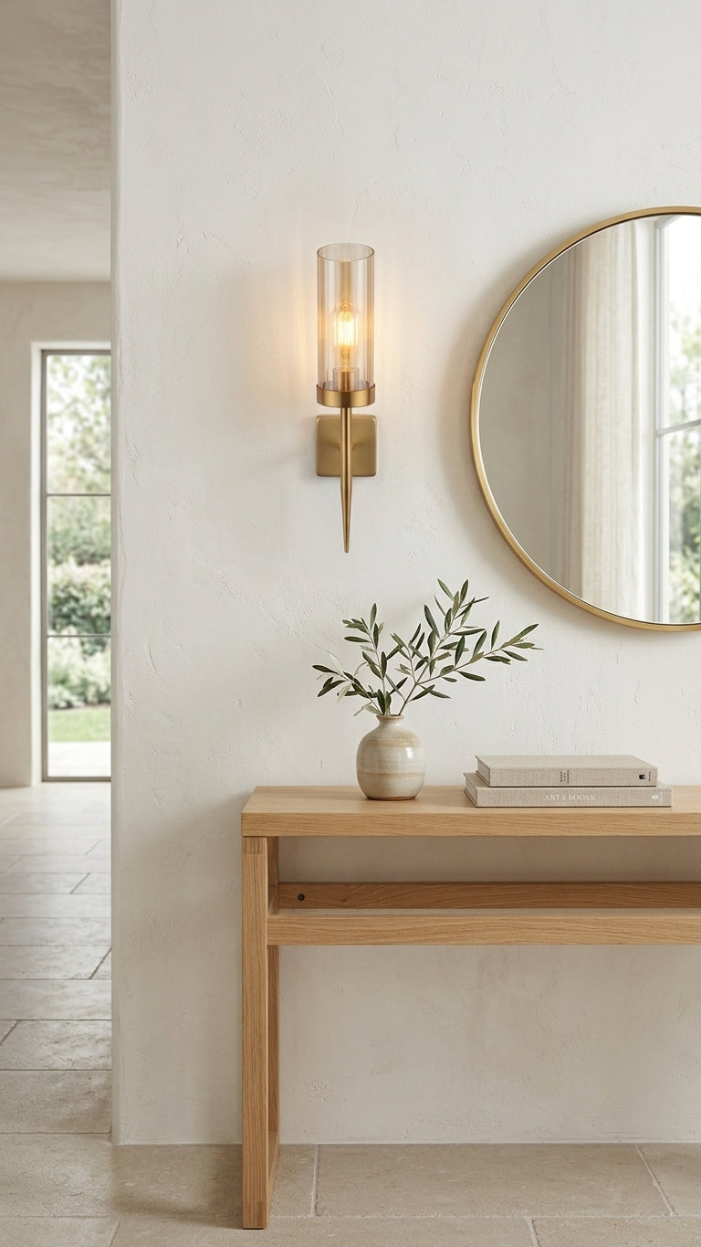 A minimalist styled entryway with a modern gold cylinder wall sconce and round mirror on a plaster wall, above a light wood console table with a ceramic vase and olive branch.