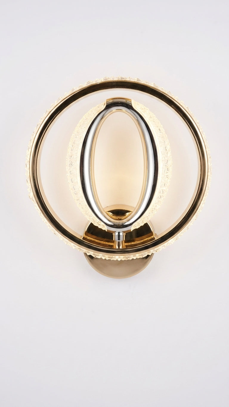 A modern wall-mounted light fixture with a circular gold-toned outer ring, a central oval glass shade, and a warm glowing light source, shown against a plain light background.