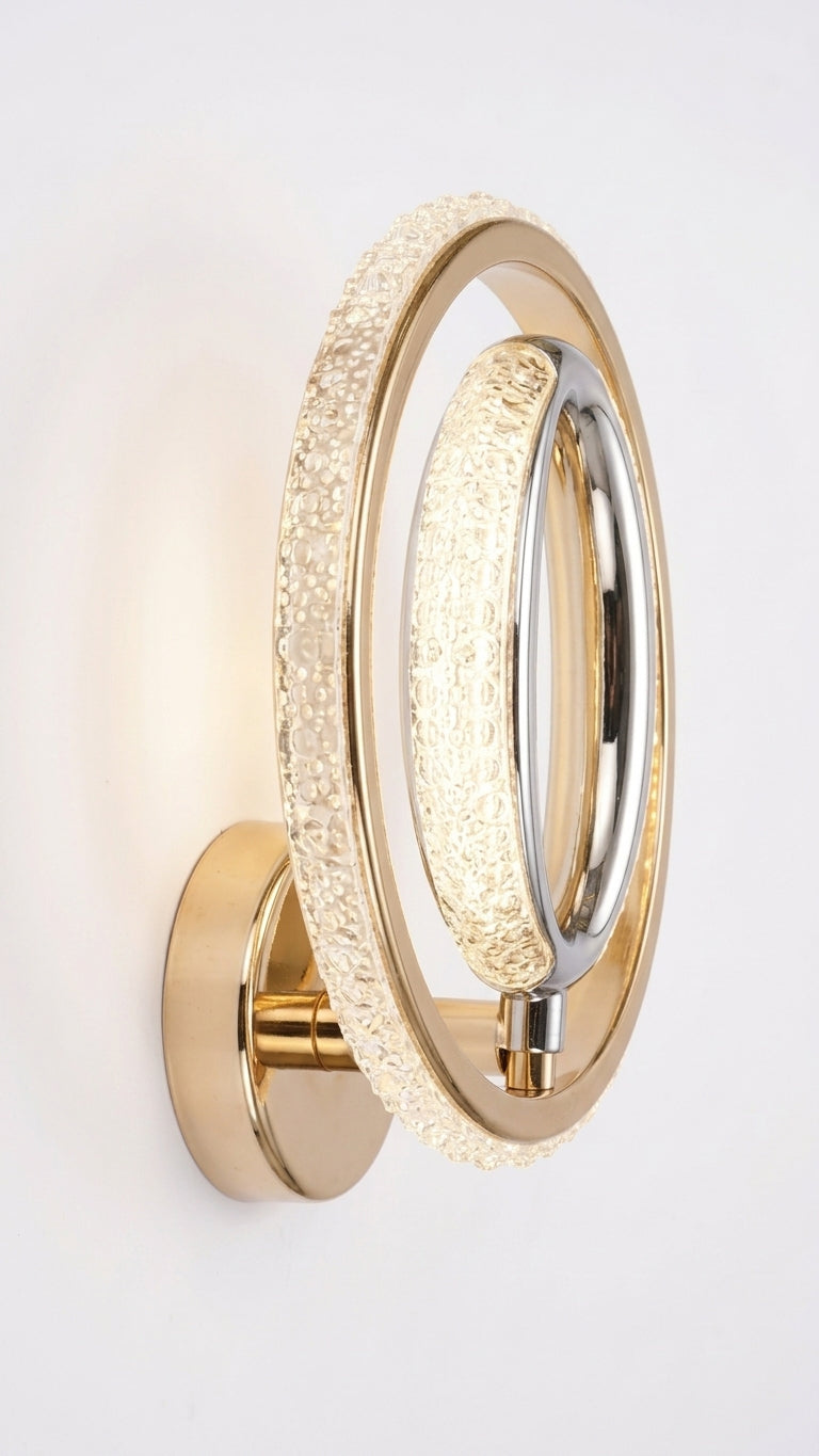 A modern wall-mounted vanity light with two concentric oval rings, one in polished gold-toned metal and the other encrusted with clear crystals that glow warm white when lit, mounted on a plain white wall.